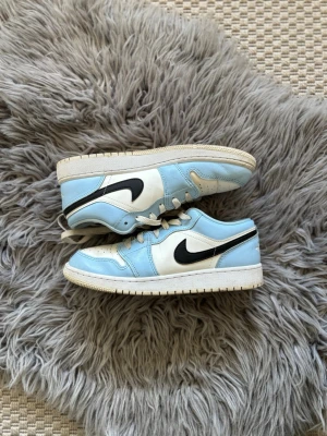 Jordan 1 Low UNC blå och vita sneakers - Snygga Jordan 1 Low sneakers i UNC-färg med ljusblå och vita paneler samt svart swoosh. Klassisk siluett med lågt skaft, snörning och vit sula. Ovandel i läder och textil, perfekt för dig som gillar streetwear och sportig stil.