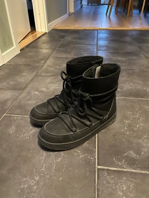 Svarta moon boots med snörning - Säljer ett par svarta moon boots med högt skaft och grov snörning runt hela skon. Bootsens ovandel är i mockaliknande material och insidan ser ut att vara fodrad för extra värme. Yttersulan är i gummi och de har dragkedja på sidan för enkel påtagning.