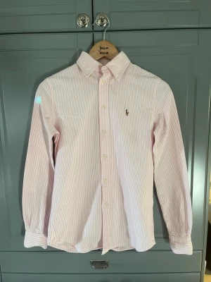 Rosa randig skjorta från Ralph Lauren - Klassisk rosa och vit randig skjorta från Ralph Lauren med lång ärm och knappar framtill. Skjortan har en broderad logga på bröstet och är tillverkad i bomull. Perfekt för en stilren och fräsch look.