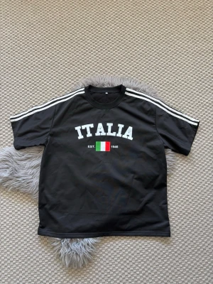 Svart t-shirt med ITALIA-tryck - Svart t-shirt med vita ränder på axlarna och stort ITALIA-tryck framtill samt italienska flaggan och texten 'EST. 1946'. Klassisk rund hals och korta ärmar. Perfekt för dig som gillar sportig stil och vill visa kärlek till Italien.