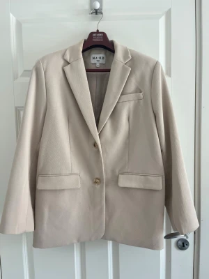 Beige kavaj från NA-KD - Stilren beige kavaj från NA-KD med klassisk passform, två knappar framtill och tre fickor. Kavajen har långa ärmar med knappdetaljer vid ärmslut och är tillverkad i ett mjukt tyg som ger en snygg look.