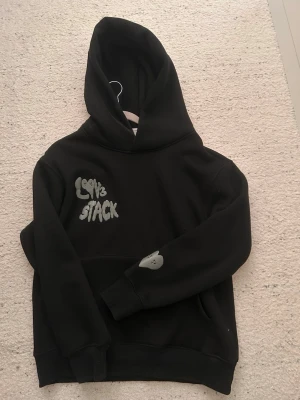 Lov3 stack - Svart hoodie med stor huva och trycket 'Lov3 Stack' i vitt på bröstet. På ena ärmen finns ett litet hjärta i samma stil. Hoodien har en klassisk passform och är gjord i mjukt material, perfekt för en chill och avslappnad look. Denna tröjan finns det inte många av 