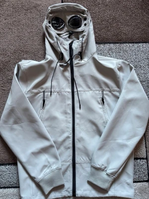 Beige vindjacka med huva - Snygg beige C.P Company vindjacka med hel dragkedja framtill och justerbar huva med snören. Jackan har två fickor med dragkedja och resår vid ärmslut och nederkant. Perfekt för blåsiga dagar och enkel att matcha med olika outfits.