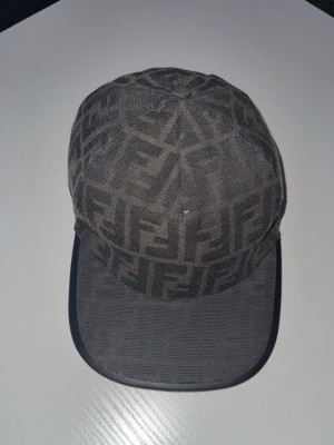 Svart fendi keps  - Snygg svart keps  med det klassiska FF-monogrammet över hela kepsen. Kepsen har böjd skärm och är tillverkad i tyg med en exklusiv känsla. Perfekt för dig som vill sticka ut med en trendig accessoar.