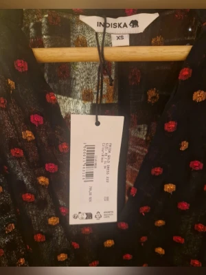 Svart prickig långklänning Indiska XS - Säljer en fantastiskt fin och festlig långärmad klänning från Indiska. Klänningen är helt ny och i absolut toppskick.   Färg & Mönster: Basen är klassiskt svart, prydd med ett dekorativt prickigt mönster i skimrande metallic-toner av guld och rosa.  Skick: Helt ny (oanvänd).  Storlek: XS  Design: V-ringad med eleganta knappar framtill och skira, långa ärmar med resår vid handlederna.  Prisinformation:  Nypris: Köpt för 799 kr   