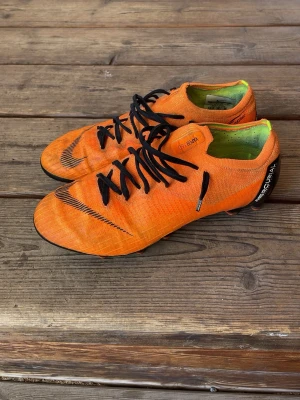 Nike Mercurial orange fotbollsskor - Säljer ett par Nike Mercurial fotbollsskor i en riktigt snygg orange färg med svarta detaljer och snörning. Skorna har en låg profil och är tillverkade i syntetmaterial för bästa känsla och grepp på planen. Perfekta för dig som vill sticka ut på fotbollsplanen.