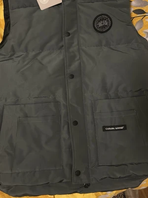 Mörkgrön dunväst från Canada Goose - Passar storlek M eller liten L. Snygg mörkgrön dunväst från Canada Goose med två stora framfickor och klassisk logotyp på bröstet. Västen har knäppning med svarta knappar och är tillverkad i slitstarkt material. Perfekt för lager-på-lager och håller dig varm under kyliga dagar.