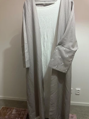 Ljusgrå lång abaya 2 delar  med vida ärmar - Lång och öppen abaya i ljusgrått med vida ärmar och dekorativa sömmar på ärmarna. Koftan har en avslappnad passform och är tillverkad i ett mjukt tyg som faller snyggt. Perfekt för lager-på-lager-stil. Underdelen är armlös och passar perfekt för sommaren.