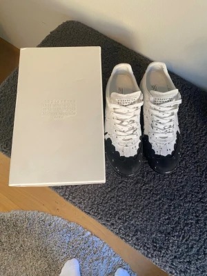 Maison Margiela svartvita sneakers - Unika sneakers från Maison Margiela i vitt tyg med svart färgstänksdesign. Låga med klassisk snörning och tydlig logga på plösen. Snygg kontrast mellan det vita och svarta, perfekt för dig som gillar statement-skor och modern streetstyle.