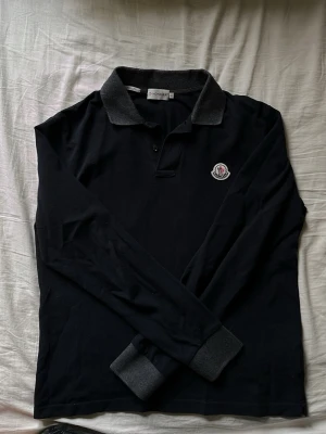 Svart långärmad Moncler henley - Snygg svart långärmad henley från Moncler med mörkgrå krage och muddar. Skjortan har en klassisk Moncler-logga på bröstet och knappar vid halsen. Perfekt för dig som gillar stilrena plagg med exklusiv känsla.