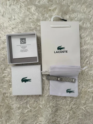 Silverfärgat armband från Lacoste - Snyggt armband i silverfärgad metall från Lacoste med rektangulära länkar och diskret Lacoste-logga. Armbandet har en stilren och modern design, levereras med originalförpackning och tygpåse med krokodiltryck.