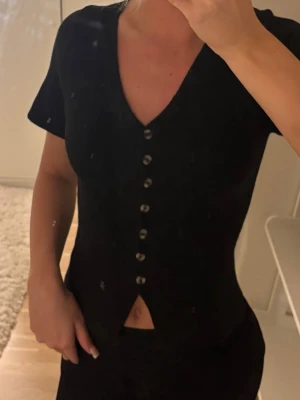 Svart topp med knappar och v-ringning - Snygg svart topp med v-ringning och dekorativa knappar hela vägen framtill. Kortärmad modell med liten slits nedtill framtill som ger en trendig touch. Perfekt att styla med både jeans och kjol för en cool look.