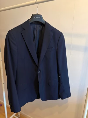 Mörkblå kavaj från Oscar Jacobson - Stilren mörkblå kavaj från Oscar Jacobson med klassisk passform och två knappar framtill. Tillverkad i exklusivt tyg från Vitale Barberis Canonico, vilket ger en lyxig känsla. Perfekt för dig som vill ha en tidlös och elegant look.   Använd endast vid två tillfällen. 