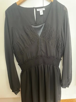 Svart långärmad klänning NLY design  - Svart klänning från NLY design  med lång ärm och v-ringad hals. Klänningen har smockdetaljer vid midjan och bröstet samt lätt transparenta ärmar. Perfekt för dig som gillar stilrena och klassiska plagg.