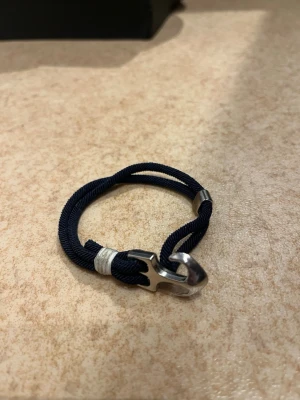 Marint armband med ankare - Snyggt marinblått armband med ett silverfärgat ankare som lås. Armbandet är gjort av ett tvinnat tygband och har detaljer i metall. Perfekt accessoar för dig som gillar nautisk stil och vill ha något unikt på handleden.