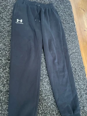Svarta mjukisbyxor Under Armour - Svarta mjukisbyxor från Under Armour med dragsko i midjan och resår vid bensluten. Byxorna har en klassisk rak passform och logga på vänster lår. Perfekta för chill eller träning. Tillverkade i mjukt material för extra komfort. Köptes på JD sports för 649kr i år (: