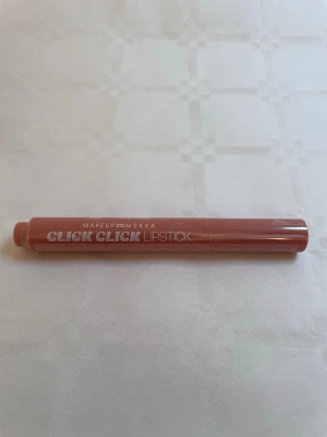 Click Click Lipstick från Makeup Mekka - Snyggt läppstift i pennform från Makeup Mekka i en mjuk nude-rosa nyans. Hylsan är smal och cylinderformad i plast med enkel klickmekanism. Perfekt för att skapa naturliga och fräscha läppar med ett modernt uttryck.