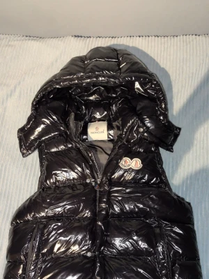 Svart glansig dunväst från Moncler - Snygg svart dunväst från Moncler med glansigt material och avtagbar huva. Västen har en klassisk Moncler-logga på bröstet och dragkedja framtill. Perfekt till sommaren och varmare väder. Den är lite sönder innuti skriv för bild men det syns inte och märks inte när man har på den på något sätt. Säljer pågrud av att jag inte andvänder den längre. Skriv för frågor och mer bilder. Storlek S men passar M.