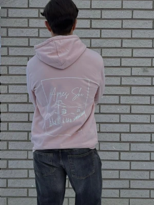 Ljusrosa hoodie med Après Ski-tryck - Ljusrosa hoodie med stor vit Après Ski-illustration på ryggen. Hoodien har en rymlig huva och är gjord i mjukt material, perfekt för att chilla efter skidåkning. Snygg och avslappnad stil som passar dig som gillar vinter och fjäll. Finns i storlekar S till XL!