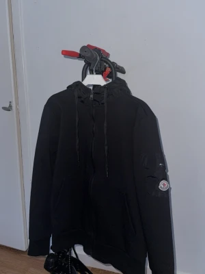 Svart moncler kofta - Svart hoodie från Moncler med dragkedja, huva och snören. På vänster ärm finns en ficka med knapp och Moncler-logga. Hoodien är tillverkad i mjukt material och har ribbade muddar vid ärmslut och nederkant. Perfekt för en avslappnad och cool look.