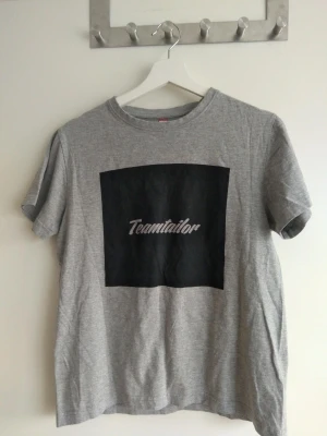Grå Teamtailor t-shirt med tryck - Grå t-shirt från Teamtailor med svart fyrkantigt tryck och vit logga på bröstet. Klassisk passform med rund hals och korta ärmar. Tillverkad i mjukt material som är skönt att bära. Enkel och stilren design som funkar till det mesta.