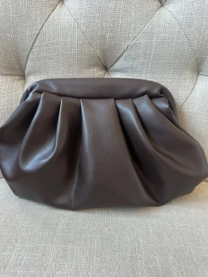 Mörkbrun clutchväska i skinnimitation - Snygg mörkbrun clutchväska i skinnimitation med mjuka veck och metallstängning. Väskan har en rymlig öppning och svart tygfoder inuti. Perfekt accessoar för att lyfta din outfit med en trendig touch. Väskan är handgjord 