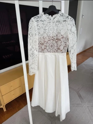 Vit spetsklänning med lång ärm från Self-Potrait - Superfin vit klänning med långärmad överdel i blommig spets och rund halsringning. Kjolen är enkel och slät i ett mjukt tyg. Perfekt för dig som vill ha en romantisk och elegant look.