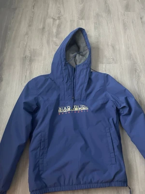 vindjacka Napapijri - Snygg blå vindjacka från Napapijri med huva och dragkedja vid halsen. Jackan har stor logga på bröstet, praktisk magficka och elastisk nederkant. Perfekt för blåsiga dagar och streetwear-stil.