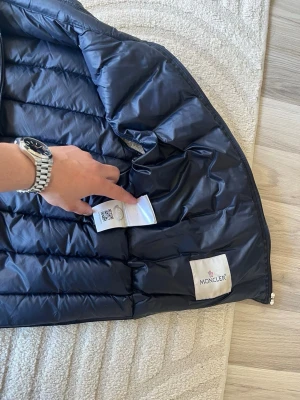 Mörkblå dunväst från Moncler - Snygg mörkblå dunväst från Moncler med klassisk quiltad design och hög krage. Västen har två dragkedjeförsedda fickor framtill och ikonisk Moncler-logga på bröstet. Tillverkad i glansigt material med dunfyllning för extra värme.