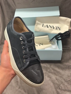 Lanvin svarta sneakers med lackad tå - Snygga svarta sneakers från Lanvin med ovandel i mocka och en glansig, lackad tå. Skorna har vita sulor, svarta skosnören och silverfärgade öljetter. Perfekta för dig som vill ha en stilren och exklusiv look.