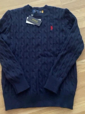 Marinblå Ralph Lauren tröja - Kabelstickad mörkblå tröja från Polo Ralph Lauren med rund halsringning och den klassiska röda logotypen broderad på bröstet. Tröjan har långa ärmar och ribbade muddar vid ärmslut och nederkant. Perfekt för en stilren och avslappnad look.