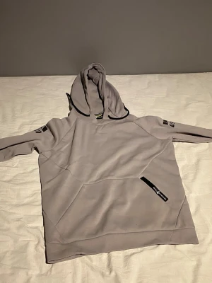 Ljusgrå hoodie med svarta detaljer - Snygg ljusgrå hoodie från Björn Borg med svart dragkedjedetalj på fickan och svarta loggor på ärmarna. Hoodien har en stor huva och en praktisk magficka framtill. Perfekt för en avslappnad och sportig stil.