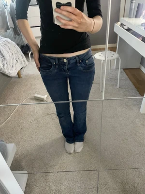 Mörkblå bootcut jeans med låg midja - Säljer ett par mörkblå jeans med bootcut-modell och låg midja. Jeansen har klassiska fem fickor, knapp- och dragkedjestängning samt snygga söm-detaljer. Perfekta för en avslappnad och trendig look.