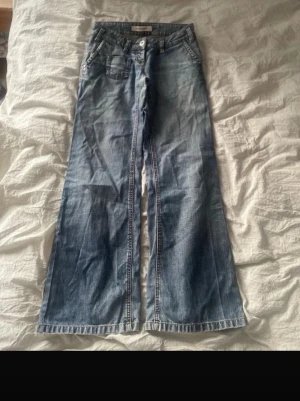 Blåa bootcut jeans med broderi - Snygga blå bootcut jeans med broderade detaljer på bakfickorna. Lite trasiga men inget som störs eller syns. Jeansen har klassisk femficksdesign, låg midja och vida ben. Perfekta för dig som gillar en avslappnad och trendig look. Tillverkade i denim med lätt tvättad effekt. Vintage 