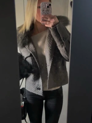 meotine jacka - Säljer en snygg grå kappa med kort modell och stor pälskrage. Kappan har dubbelknäppning framtill och är tillverkad i ett strukturerat tyg som ger en exklusiv känsla. Perfekt för dig som vill ha en trendig och varm jacka.