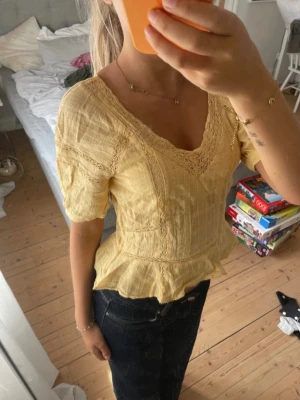 Gul blus med spetsdetaljer - Söt gul blus med v-ringning och spetsdetaljer. Blusen har korta ärmar, markerad midja och volang nertill. Tillverkad i ett lätt och luftigt material som passar perfekt till varmare dagar.