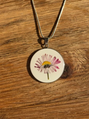 Halsband med blomma i rund berlock - Snyggt halsband med en rund vit berlock med en tusensköna.. Kedjan är silverfärgad och tunn, vilket ger ett stilrent intryck. Perfekt accessoar för dig som gillar naturinspirerade smycken och vill ha något unikt.