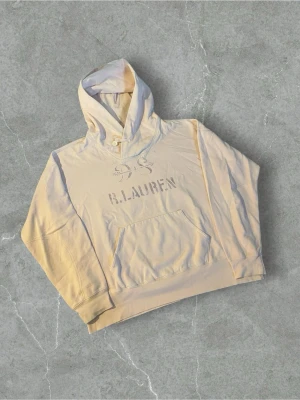 Rosa hoodie från Ralph Lauren Denim & Supply | XL - Snygg beige hoodie från R. Lauren med tryckt logga och text på bröstet. Hoodien har en stor huva med knappdetalj och en klassisk magficka framtill. Perfekt för en avslappnad och trendig stil.