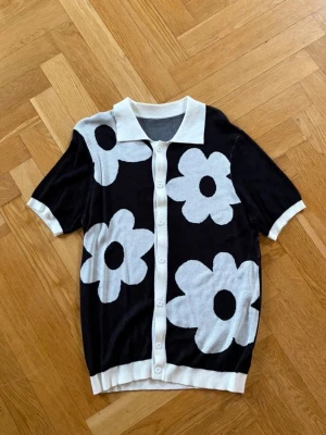 Svart kofta med stora blommor - Kortärmad svart kofta med stora vita blommor och vita detaljer vid krage, knappslå, ärmslut och nederkant. Koftan har en klassisk krage och knäpps framtill med vita knappar. Perfekt för dig som gillar retro och lekfull stil.