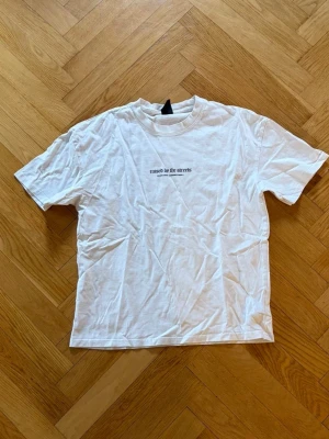 Vit t-shirt med tryck - Vit t-shirt med svart texttryck 'raised on the streets' på bröstet. Klassisk rund halsringning och korta ärmar. Perfekt basic-plagg med enkel och clean stil, passar till allt.