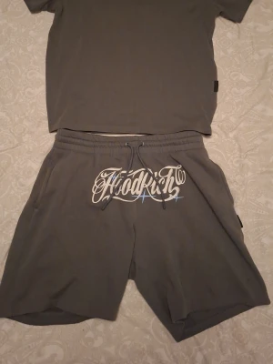 Grått set från Hoodrich med tryck - Säljer ett grått set från Hoodrich med t-shirt och shorts. Båda delarna har vit tryckt logga och text framtill. T-shirten har rund hals och korta ärmar, medan shortsen har resår och snörning i midjan. Perfekt för en avslappnad streetwear-look.