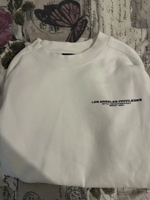 Vit loose fit hoodie från H&M - Säljer en vit hoodie från H&M i loose fit-modell. Tryck på bröstet med texten 'Los Angeles Privileges'. Mjuk insida och ribbade muddar vid ärmslut och nederkant. Perfekt för en avslappnad och trendig stil.