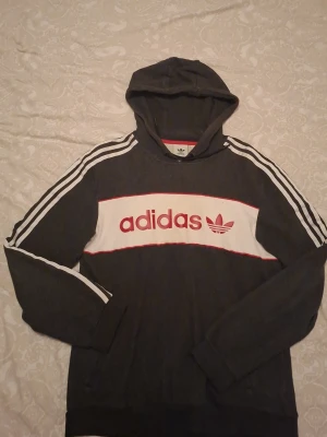 Adidas mörkgrå hoodie med ränder - Mörkgrå hoodie från Adidas med klassiska vita ränder längs ärmarna och en vit panel med röd Adidas-logga på bröstet. Tröjan har huva och långa ärmar, samt ribbade muddar. Perfekt för dig som gillar sportig och avslappnad stil.
