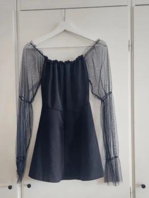 Svart kort klänning med meshärmar - Trendig svart kort klänning med offshoulder-design och långa meshärmar. Klänningen har en lätt utställd kjol och transparenta ärmar som ger en cool och modern look. Perfekt för dig som vill sticka ut med en unik stil.
