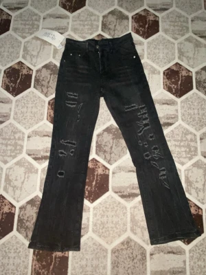 Svarta bootcut jeans med slitningar - Säljer ett par svarta bootcut jeans med coola slitningar och råa detaljer på framsidan. Jeansen har hög midja, klassisk femficksmodell och är tillverkade i denim. Perfekta för dig som gillar en edgy och trendig stil.