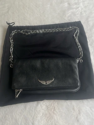 Svart axelväska med kedja  - Snygg svart axelväska i skinnimitation med silverfärgad kedjerem och ett metallmärke framtill. Väskan har en stilren design med dragkedja och magnetknapp. Perfekt för dig som gillar en edgy och trendig look.
