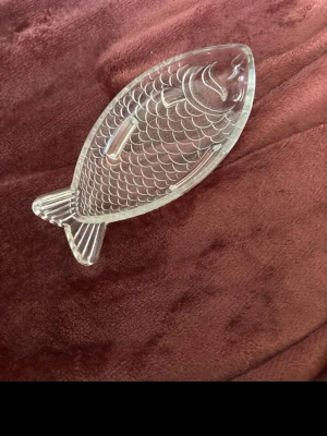 Glasfat i form av fisk - Säljer ett genomskinligt glasfat format som en fisk. Fatet har detaljerade fiskfjäll och fenor ingraverade i glaset, vilket ger en unik och dekorativ look. Perfekt som serveringsfat eller dekoration i hemmet.