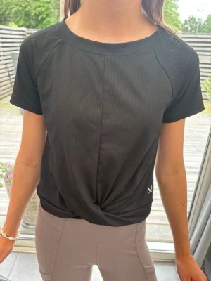 Svart sportig t-shirt med twist - Modellen är 157. Svart sport-tshirt med kort ärm och twistad detalj framtill. Tillverkad i ett lätt och stretchigt material som andas, perfekt för träning eller gym. Diskret logga nertill på sidan. Enkel och stilren design med rund halsringning.