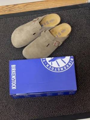 Beige mockatofflor från Birkenstock - Säljer ett par klassiska Birkenstock tofflor(bostons) i beige mocka med justerbart spänne i metall. Modellen har en öppen häl och mjuk innersula för extra komfort. Perfekta för dig som gillar stilrena och bekväma skor.
