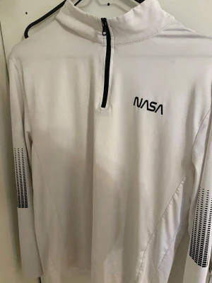 Vit långärmad NASA polotopp - Säljer en vit långärmad polotopp med svart NASA-tryck på bröstet och coola svarta detaljer på ärmarna. Toppen har en hög krage och half zip framtill. Perfekt för dig som gillar rymdtema och sportig stil.